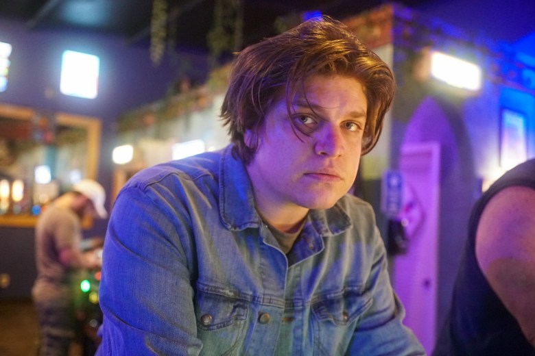 An indoor portrait photograph showing musician Anthony Santaniello of Antro Banks. Captured in a medium close-up shot inside a dimly lit venue, Santaniello leans forward on a counter while looking directly into the camera lens with medium-length brown hair. He wears a blue denim button-up shirt over a dark tee under vibrant purple and blue ambient bar lighting, with an archway, warm background lights, and another person visible in the blurred background.