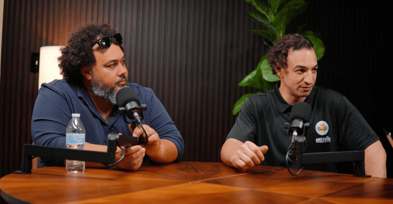 A podcast recording photograph showing Tony Adams (L) and Tyler Sirota at CLTV studio in Tampa on Sept. 10, 2026. Captured in a medium shot seated at a polished wooden table, Tony Adams (left) sits wearing a dark blue polo shirt, sunglasses pushed up onto his curly hair, and a dark beard, looking toward Tyler Sirota (right), who wears a black polo shirt with an "ART FAIR TAMPA" logo patch. Both men have broadcast microphones mounted on adjustable boom arms on the table in front of them, set against a dark fluted wood wall and a green leafy houseplant under warm indoor studio lighting.