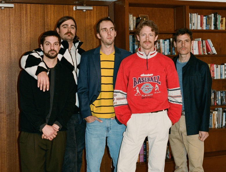 An indoor group portrait photograph by C.J. Harvey showing folk-rock band Caamp. Captured in a medium wide shot standing indoors, five band members pose together in front of wooden doors and a wooden bookcase filled with books. The member in the front center wears a bright red "BASEBALL" sweatshirt and white trousers, flanked on the left by a band member in a dark blue blazer with a yellow-and-black striped shirt and light blue jeans, and on the far left by two members in dark jackets and a patterned coat. The fifth member stands on the right in a dark jacket and khaki trousers under warm flash lighting.