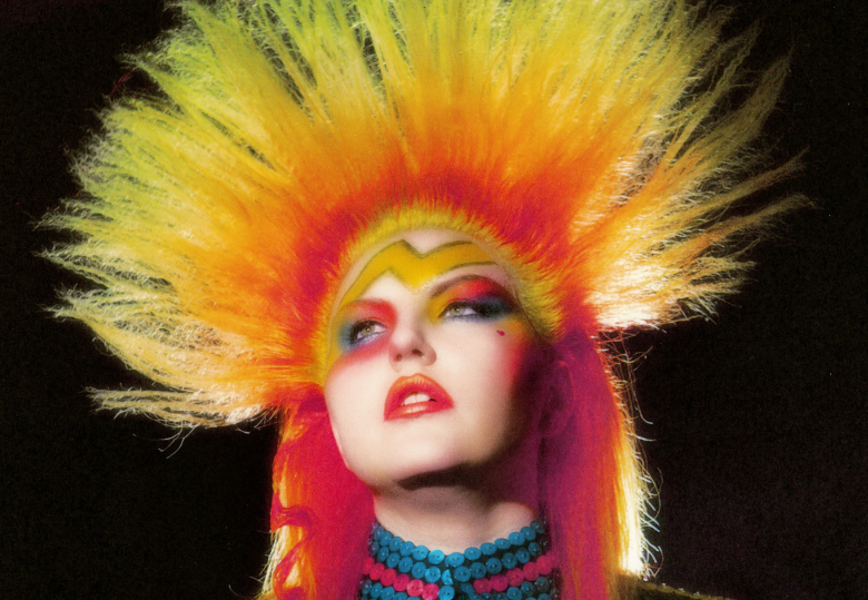 A dramatic editorial press photograph showing musician Caroline Kingsbury. Captured in a tight close-up portrait angled slightly upward against a pitch-black background, Kingsbury tilts her head back while staring forward with bold, avant-garde makeup. She features yellow-and-orange spiky teezed hair rising like a flame around her head, a yellow lightning-bolt accent painted across her forehead, bright red-and-yellow eyeshadow with bold eyeliner, vivid red lipstick, and a multi-row turquoise beaded choker necklace around her neck under intense studio lighting.