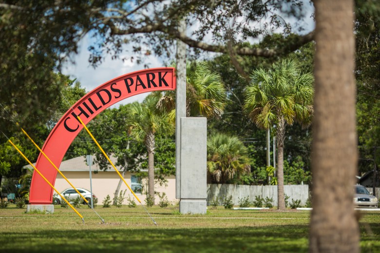 An outdoor landscape photograph showing Childs Park in St. Petersburg, Florida. Captured in a medium wide shot through a tree trunk in the foreground, a prominent curved red archway sign reading "CHILDS PARK" in capital lettering stands in a grassy park setting next to a tall, grey rectangular stone pillar monument. In the background, lush green oak trees, palm trees, a residential house, and a light-colored perimeter wall sit under a partly cloudy sky in natural daylight.
