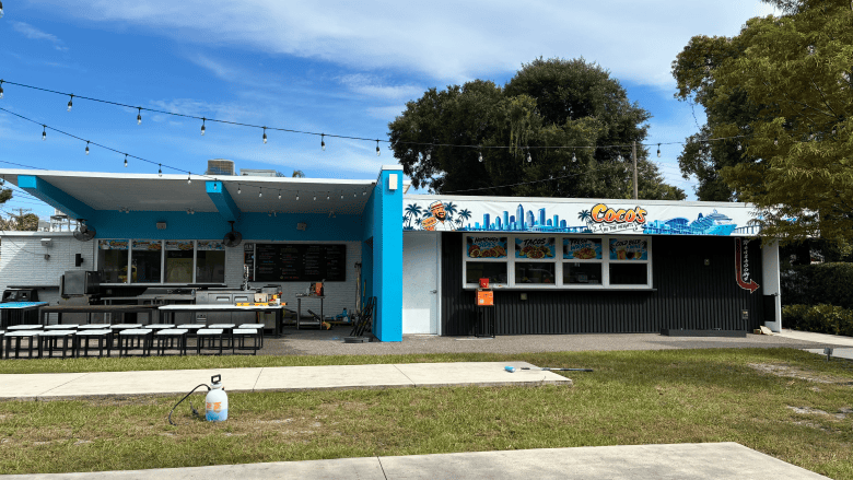 An outdoor architectural photograph by Annalise Mabe showing Coco's In the Heights in Tampa, Florida on Sept. 29, 2026. Captured in a wide shot under a clear blue sky, the blue and black single-story venue features a covered outdoor patio on the left with picnic tables, bench seating, and overhead string lights. On the right, a black ordering counter with a four-panel window is decorated above with painted murals of palm trees, ocean waves, and stylized text reading "Coco's", set against a lawn forecourt with trees in the background.