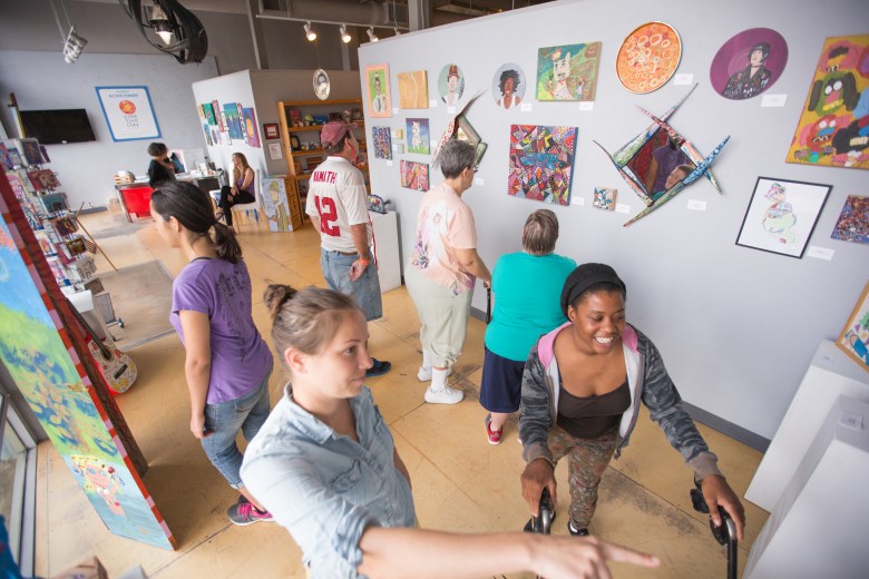 An interior gallery photograph showing Creative Clay, c. 2016. Captured from an elevated high-angle wide perspective, art gallery attendees and artists browse artwork displayed along gray walls in a brightly lit studio. In the foreground, a woman in a light blue denim shirt points toward a display on the right while a smiling woman in a hoodie stands nearby; behind them, other visitors admire colorful paintings, framed prints, and handmade crafts arranged across the gallery space under industrial track lighting.