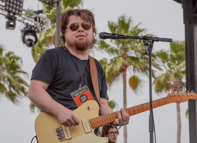 A festival concert photograph by Dan Schuman showing Damon Fowler plays Gasparilla Music Festival at Curtis Hixon Park in Tampa, Florida on March 13, 2016. Captured in a medium close-up shot, blues musician Damon Fowler plays a butterscotch blonde Telecaster-style electric guitar in front of a stage microphone stand. He wears sunglasses, a dark t-shirt, and a red GMF festival lanyard around his neck, set against an outdoor festival backdrop of palm trees and stage lighting rigs under soft natural daylighting.