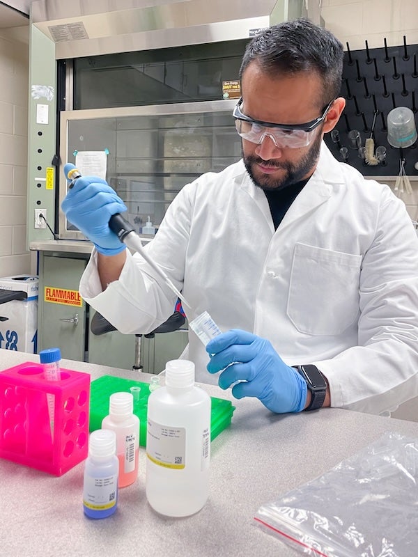 A researcher wearing a white lab coat, safety glasses and blue gloves uses a pipette to transfer liquid into a sample tube at a laboratory workbench.