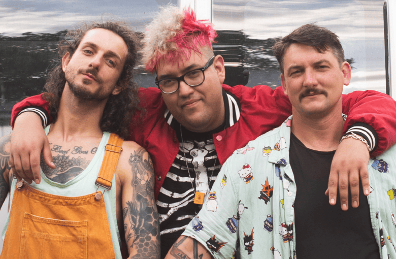A promotional press photograph showing the Tampa punk band Lychee Camp. Captured in a medium three-person group shot outdoors, the middle band member has vibrant pink-and-blonde dyed hair, glasses, a black-and-white skeleton print top, and a red varsity-style bomber jacket, draping arms around the shoulders of both bandmates. On the left, a member with shoulder-length curly dark hair and chest/arm tattoos wears orange overalls over a light tank top; on the right, a member with a mustache wears a short-sleeved light-blue patterned button-up shirt over a black t-shirt, set against a dark reflective building backdrop under bright daytime lighting.