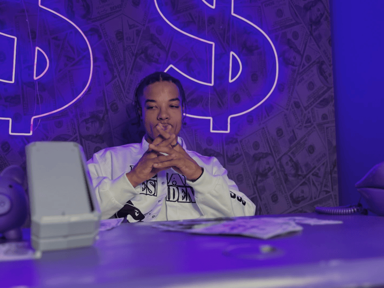 A promotional portrait photo showing MacDeezy the rapper. Captured in a medium shot, rapper MacDeezy sits behind a desk with hands clasped under his chin, looking forward. He wears a white long-sleeve sweatshirt with black text reading "PRESIDENT" alongside black graphic prints. In front of him on the desk sit stacks of cash and a money-counting machine, set against a dark wall covered in a pattern of US dollar bills and illuminated by bright purple neon dollar-sign signs ($) under dim blue and neon stage lighting.