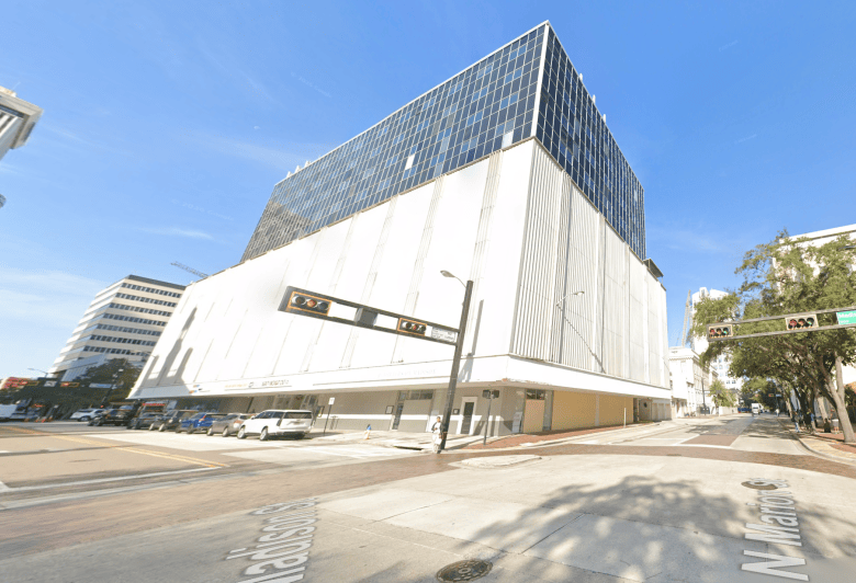 A street view screenshot from Google Maps showing The Madison building in downtown Tampa. Captured in a wide-angle corner view at the intersection of Madison Street and N. Marion Street, the multi-story commercial structure features a solid white lower façade with vertical ridges and a glass-windowed upper section. Horizontal traffic lights extend across the foreground street corner under a clear blue sky and bright daytime sunlight.