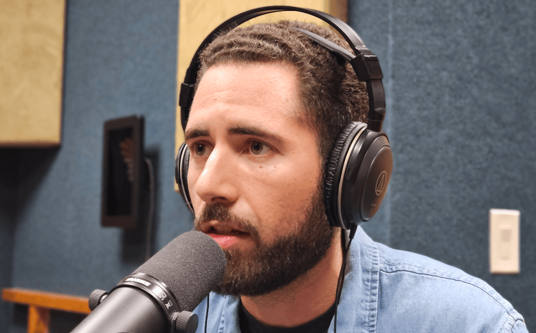 A radio studio broadcast photograph by Ray Roa showing Manny Ramirez at WMNF studios in Tampa, Florida on April 3, 2026. Captured in a close-up profile view, Manny Ramirez speaks into a black broadcast microphone with foam windscreen while wearing large black Audio-Technica over-ear headphones. He features short braided hair, a full dark beard, and a light blue denim shirt over a black t-shirt, set against blue soundproof acoustic wall panels and studio acoustic treatment tiles under bright interior lighting.