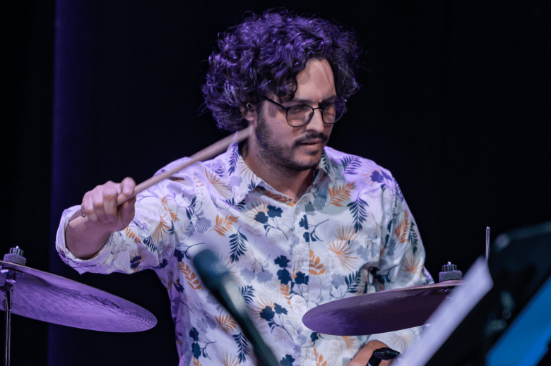 A concert performance action photograph by Stephen Splane showing Mark Feinman of La Lucha at Hillsborough Community College's Mainstage Theatre in Ybor City, Florida on Sept. 11, 2022. Captured in a medium shot behind a drum kit, drummer Mark Feinman strikes a ride cymbal with a drumstick while focused intently on his performance. He wears dark-rimmed glasses, dark curly hair, and a white short-sleeved button-up shirt patterned with blue and gold tropical leaves, framed by cymbals and music stands under purple and blue stage lighting.