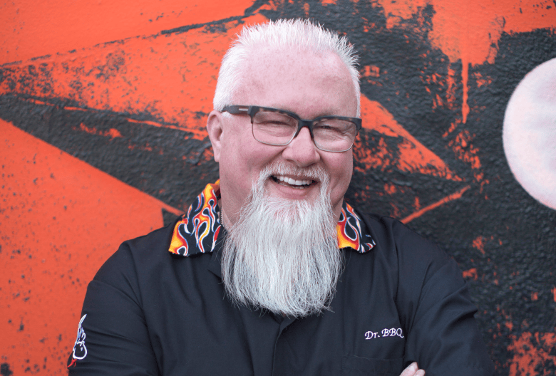 A promotional portrait photograph showing pitmaster Ray Lampe, aka Dr. BBQ, c. 2017. Captured in a medium close-up shot, Ray Lampe smiles directly at the camera with framed eyeglasses, white hair, and a full white beard. He wears a black short-sleeved chef jacket featuring a colorful flame-patterned collar, set against a red and black graphic wall backdrop.