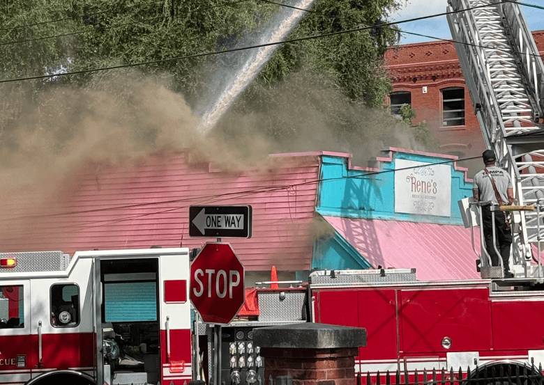 An emergency news photograph by Tiffany Haas showing the Tampa Fire Department works on a fire at Rene's Mexican Kitchen in Tampa, Florida on Sept. 30, 2026. Captured in a daylight shot from street level, smoke rises above the pink-and-teal wooden exterior of Rene's Mexican Kitchen as a high-pressure water stream sprays onto the roof. A red fire truck rescue vehicle, a ladder truck with a firefighter positioned high on the aerial platform, and prominent street signage including a red octagon "STOP" sign and a black "ONE WAY" arrow sign stand in the foreground.