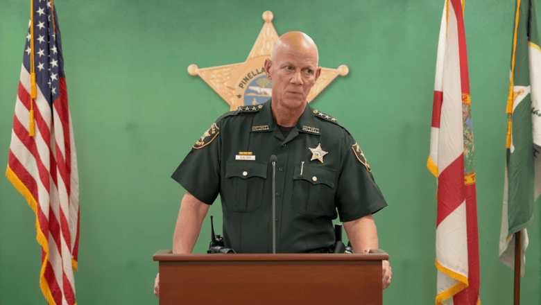 A screengrab from a Sept. 28, 2026 press conference led by Pinellas Sheriff Bob Gualtieri. Captured in a medium shot behind a dark wooden podium, Pinellas County Sheriff Bob Gualtieri stands wearing a dark law enforcement uniform with shoulder patches, a name badge, and a silver star emblem. He stands centered before a green backdrop featuring a large gold sheriff's star logo, flanked by the American flag on the left and the Florida state flag on the right under indoor press room lighting.