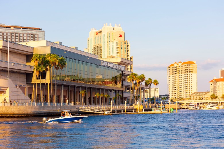 A waterfront architectural photograph by Keir Magoulas showing Tampa Convention Center in Tampa, Florida on July 3, 2023. Captured in a wide horizontal shot across the Garrison Channel, a small blue-and-white motorboat cruises along the water in front of the modern multi-story glass-and-masonry Tampa Convention Center facade. Tall palm trees line the waterfront walkway, with high-rise hotels and city buildings set against a clear sky illuminated by warm late-afternoon sunlight.