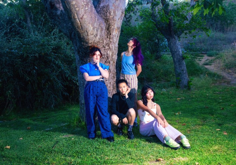 A promotional press photograph by Jaxon Whittington showing The Linda Lindas. Captured in a vertical shot on a grassy park lawn under a large tree trunk, the four members of LA punk band The Linda Lindas pose together in colorful casual attire—including bright blue tops, dark sweaters, light pants, and green sneakers—with two members standing against the trunk and two seated on the grass under soft outdoor daylighting.