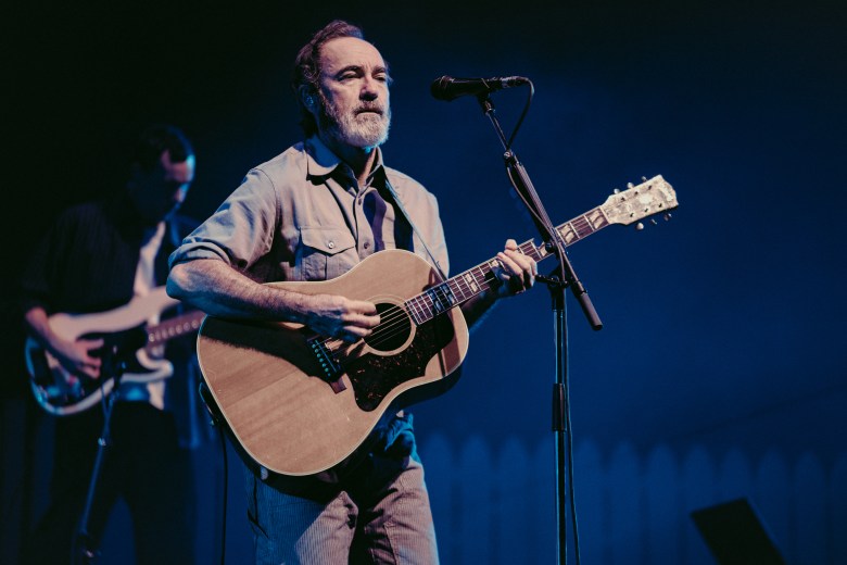 A concert performance photograph by Ryan Metcalf showing The Shins play Benchmark International Arena in Tampa, Florida on Oct. 14, 2026. Captured in a medium shot under cool blue stage lighting, front man James Mercer stands center stage singing into a stand-mounted microphone while strumming a natural wood acoustic guitar. He wears a gray short-sleeved button-up work shirt with dual chest pockets and has a trimmed gray beard, with a background musician playing electric guitar in soft focus against a dark stage backdrop.