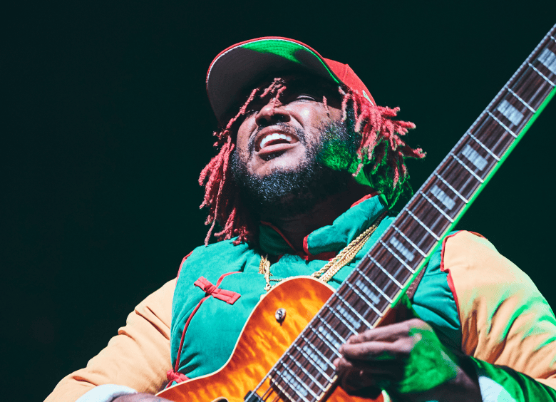 A concert performance action photograph by Koury Angelo showing musician Thundercat plays Hollywood Forever Cemetery as part of Red Bull Music Academy in Los Angeles, California on Oct. 14, 2017. Captured in a low-angle shot on a dark stage, bassist Thundercat looks upward with an open-mouthed expression while playing a custom 6-string Ibanez bass guitar with a sunburst flame-maple finish. He wears red dreadlocks, a red-and-green baseball cap, a gold chain necklace, and a green-and-yellow long-sleeved jacket with red piping under green stage spotlighting.