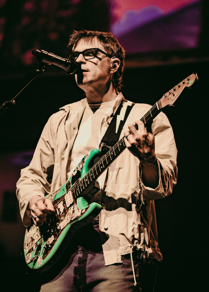 A concert performance photograph by Ryan Metcalf showing Weezer plays Benchmark International Arena in Tampa, Florida on Oct. 14, 2026. Captured in a medium three-quarter shot under warm stage lighting, Weezer frontman Rivers Cuomo stands singing into a stand-mounted microphone while playing a sticker-covered seafoam green electric guitar. He wears thick black-rimmed glasses, a beige zip-up jacket over a white t-shirt, dark jeans, and a black guitar strap with a white lightning bolt graphic against a dark background with pink ambient illumination.