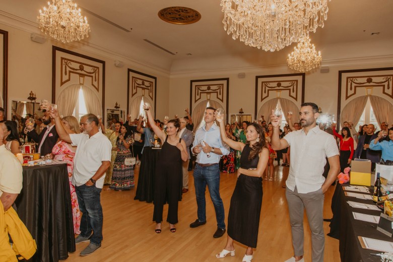 A wide interior event photograph by Centro Asturiano de Tampa showing Wines of Spain at the Centro Asturiano de Tampa in Ybor City, Florida on Oct. 16, 2024. Captured from a wide angle across the polished hardwood floor of a grand ballroom, attendees stand raising wine glasses in a collective toast. In the foreground, guest figures stand beside high-top cocktail tables and tasting stations beneath crystal chandeliers and grand arched wall moldings under warm venue lighting.