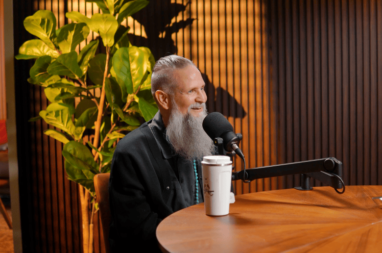 A podcast interview photograph showing artist Chris Parks, aka Palehorse, at Creative Loafing Tampa Bay's CLTV studio in Tampa, Florida on Sept. 24, 2026. Seated profile-view at a wooden round table in a podcast studio, artist Chris Parks smiles with a long grey beard, hair styled in a high fade topknot, and a dark jacket while speaking into a desk-mounted broadcast microphone. A white travel coffee mug sits on the table in front of him, set against a dark slatted wooden accent wall and a potted leafy green plant under warm indoor studio lighting.