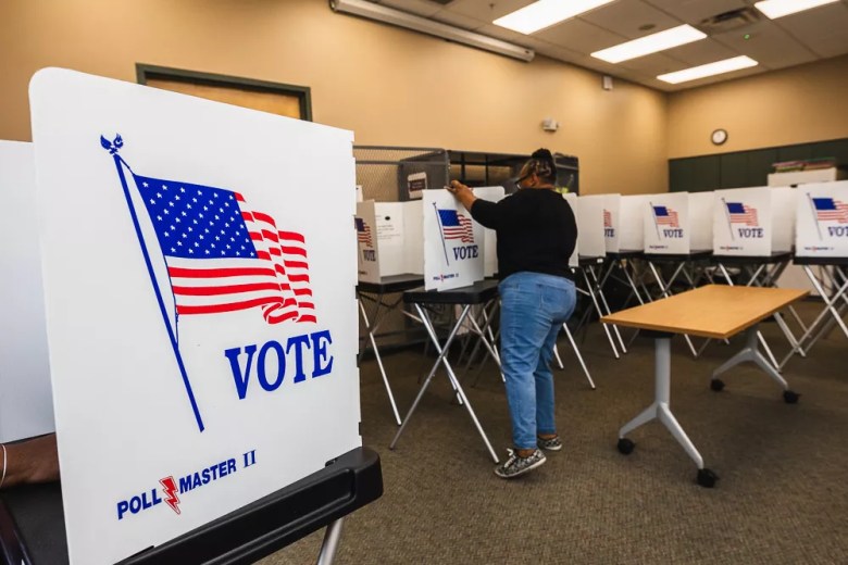 A news photograph showing Hillsborough County's Supervisor of Elections office tests equipment in Tampa, Florida on July 18, 2024. Captured in a wide indoor shot inside a voting facility, a voter wearing a dark shirt and blue jeans stands in the middle ground filling out a ballot at a voting booth, surrounded by rows of white "VOTE" privacy screens and voting stations set up across a carpeted room under overhead fluorescent panel lights, with a close-up privacy screen labeled "POLLMASTER II" displaying an American flag and "VOTE" graphics in the immediate foreground.