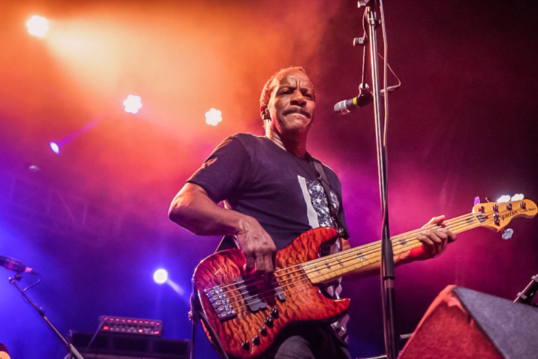 A concert performance photograph showing Dumpstaphunk plays Jannus Live in St. Petersburg, Florida on Sept. 28, 2017. Captured in a low-angle medium shot, bassist Tony Hall stands on stage playing a flame maple top electric bass guitar while positioned beside a microphone stand. He wears a dark t-shirt featuring a stylized flag motif, performing under vivid orange, red, and purple stage spotlights with bright overhead lighting fixtures illuminating the dark background.