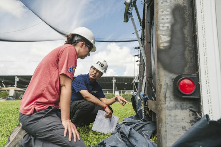Two people in hard hats examine the bottom of a tipped-over mobile home.