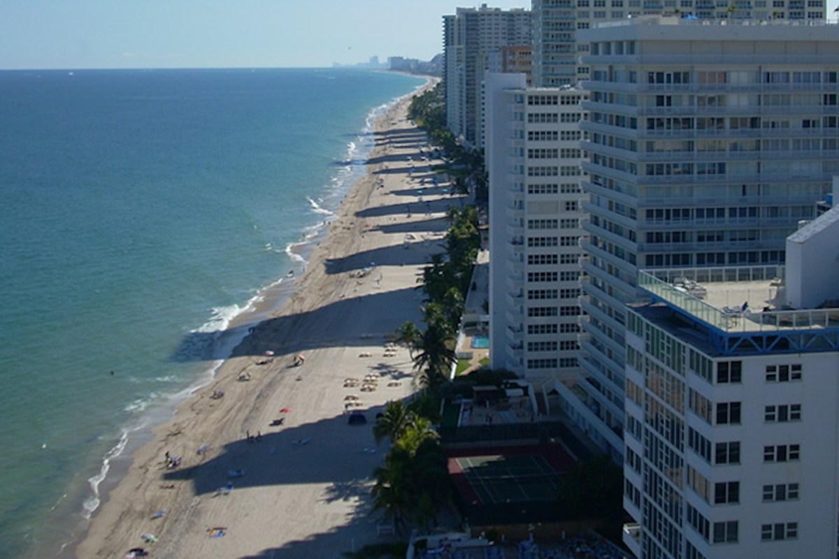 A view of a beach lined with buildings, featuring palm trees and sunbathers along the shoreline.