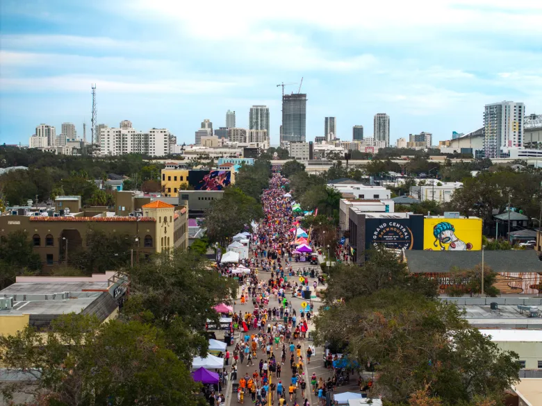 An elevated drone photo showing Halloween On Central in St. Petersburg, Florida on Oct. 27, 2024. Captured looking down Central Avenue in the Grand Central District, thousands of attendees fill the pedestrian-only street lined with vendor tents, colorful murals, and trees. The city skyline and surrounding buildings extend into the distance under a partly cloudy sky illuminated by soft daylighting.