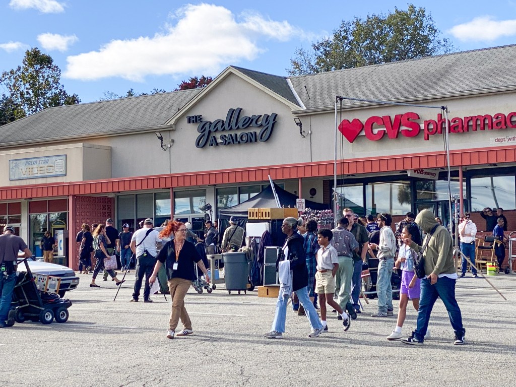 Behind-the-scenes view of the "Miami Vice '85" film set in a New Jersey strip mall, with crew and background actors milling about.