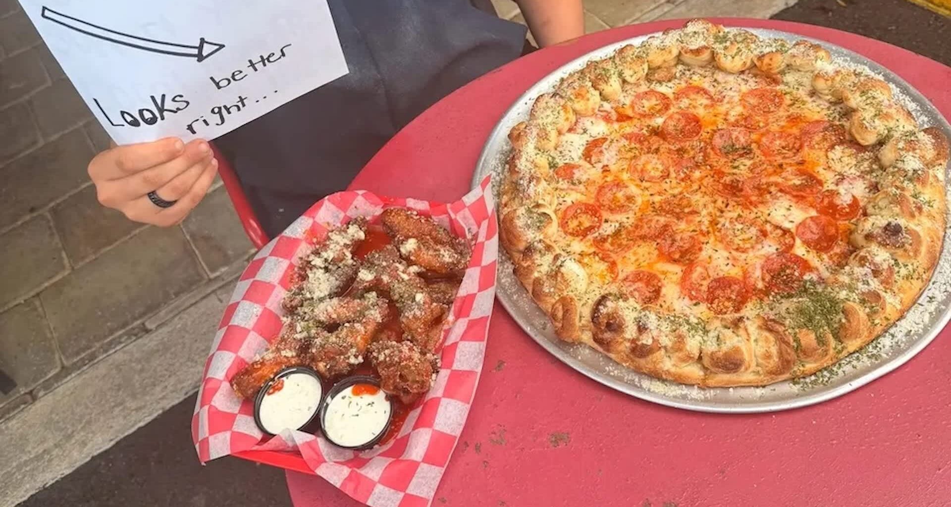 A pizza with pepperoni and a basket of chicken wings with dipping sauces on a red table.