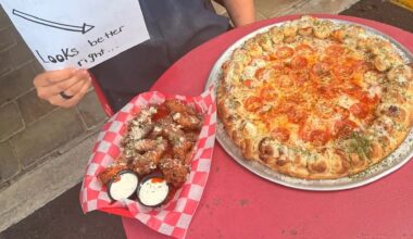 A pizza with pepperoni and a basket of chicken wings with dipping sauces on a red table.