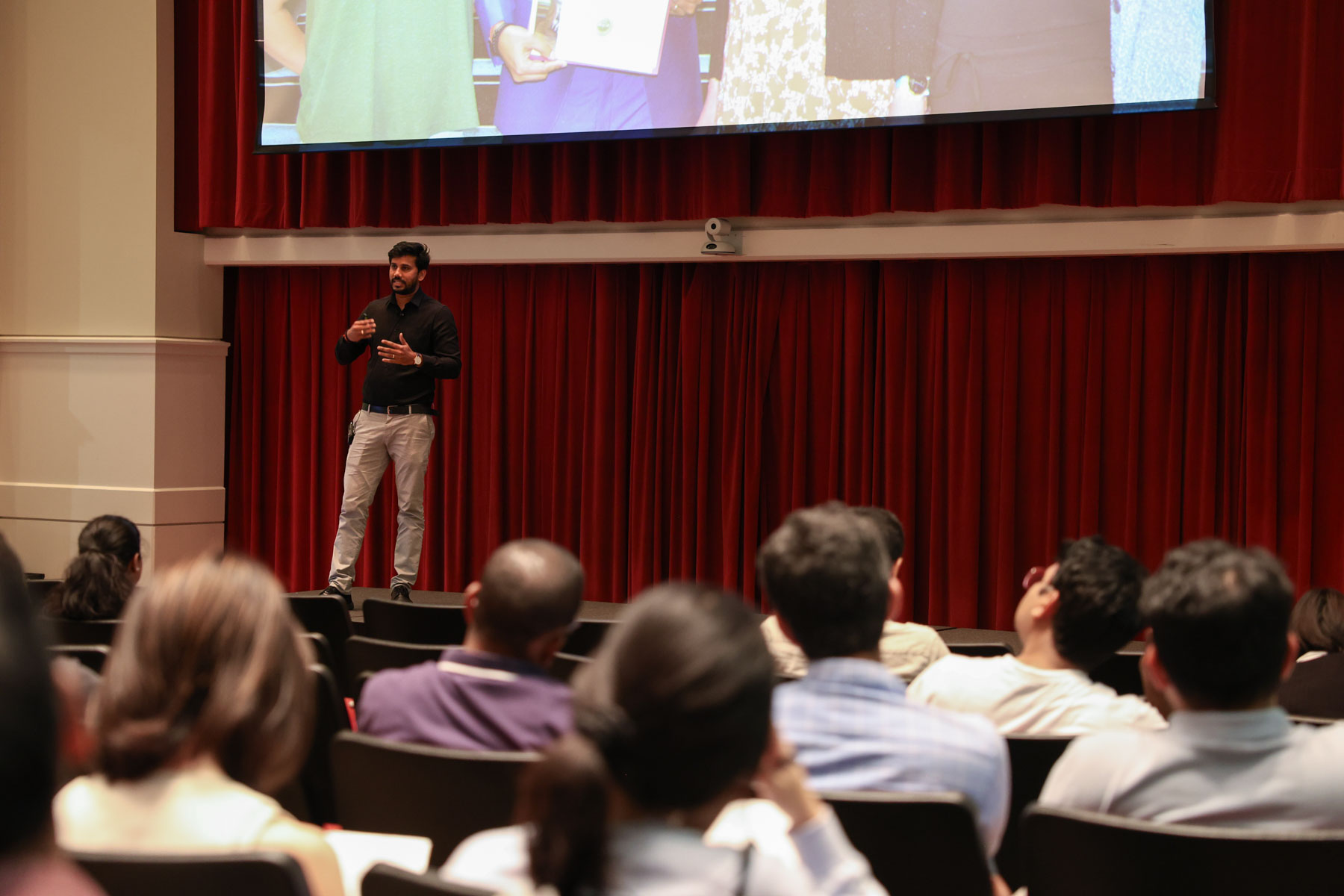 A man speaks on a stage in front of a red curtain and a large screen while the audience watches from auditorium seating.
