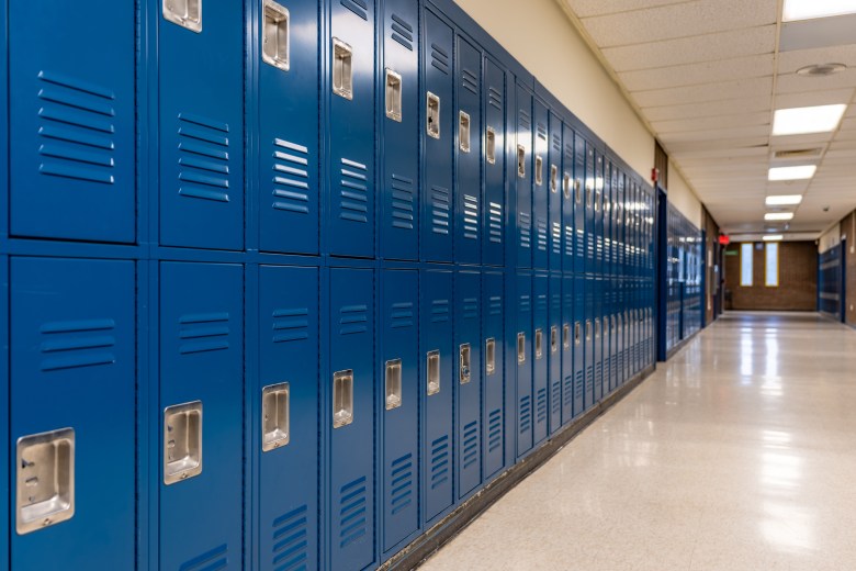 Row of blue metal student lockers with built-in combination locks along a brightly lit, empty school hallway.