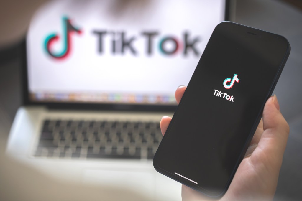 A person holding a smartphone displaying the TikTok app logo, with a laptop showing the TikTok logo in the background.
