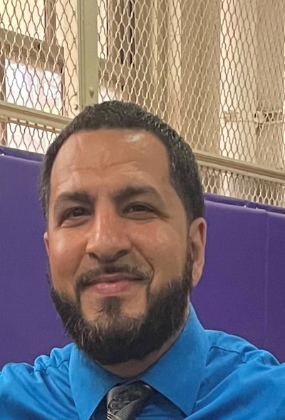 A photograph of a man wearing a blue dress shirt posing for a portrait with a window in the background.