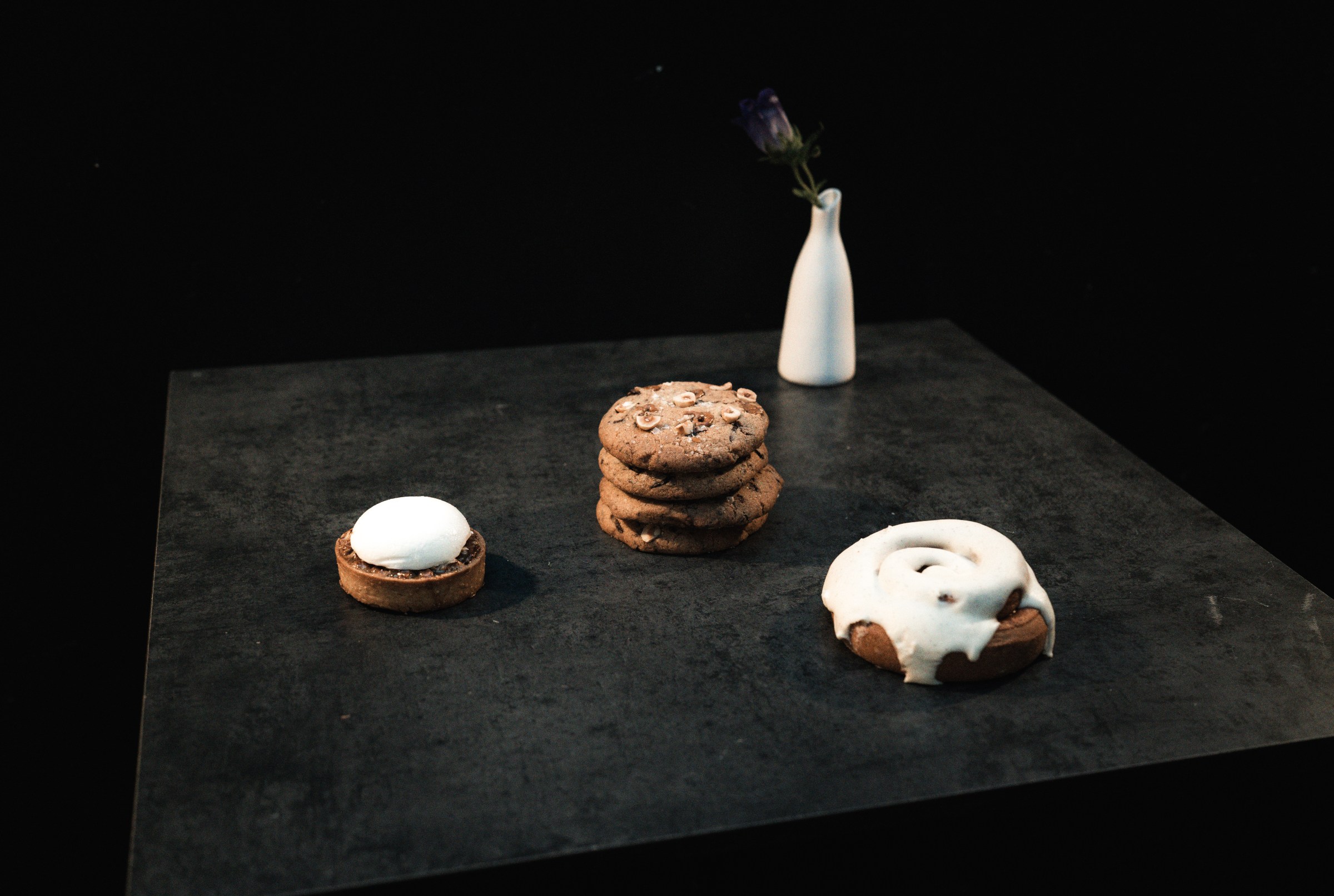 A dark table with a tart, a pile of cookies, and a cinnamon bun.