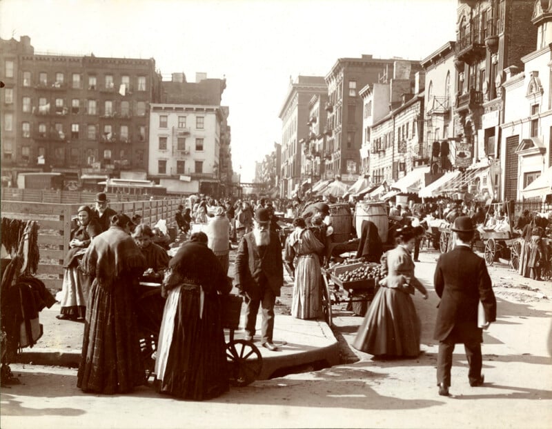 A sepia-toned photo of a busy city street from the early 1900s shows people in period clothing walking, shopping at vendor carts, and socializing among tall buildings and market stalls.