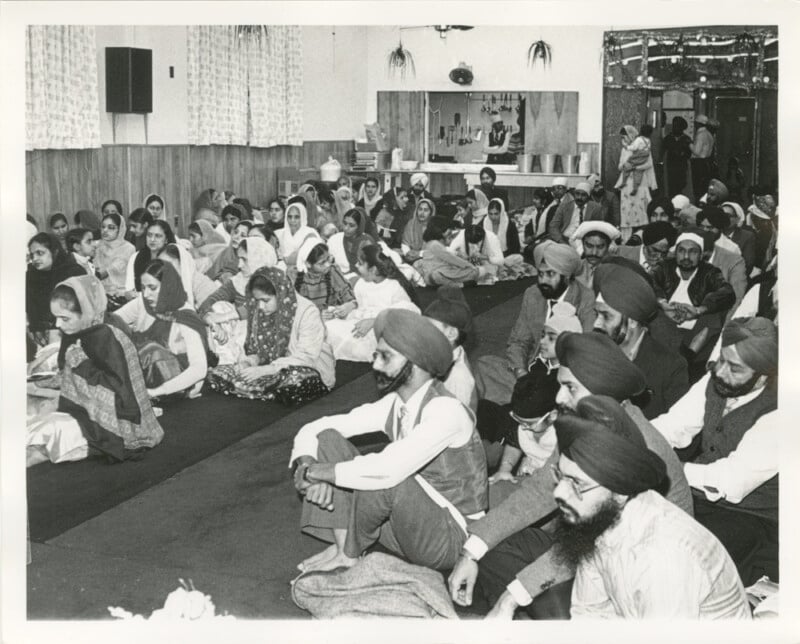 A group of Sikh men, women, and children sit cross-legged on the floor of a gurdwara, attentively listening. Most are wearing turbans or head coverings. The room has simple decor with curtains and framed photos.