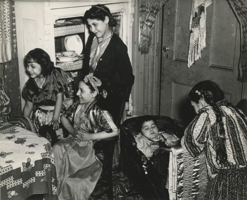 Five children in patterned clothing gather in a cozy room; one boy reclines in a stroller while the others, mostly girls, stand or sit nearby, smiling or looking down. The setting is homely, with a table, fireplace, and dishes visible.