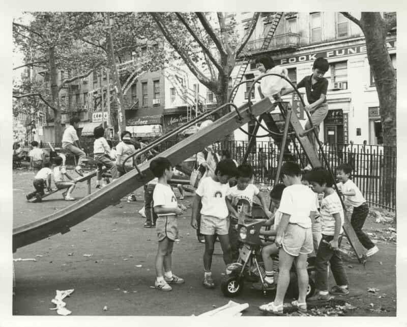 Black-and-white photo of children playing in a city playground. Some kids climb and slide, others gather around a tricycle. Trees and buildings are in the background, with adults and more children nearby.