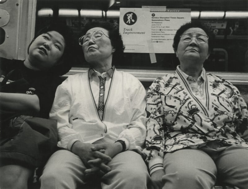 Three older women with glasses sit closely together on a subway bench, with neutral or tired expressions. A "Track Improvement" notice is posted on the wall behind them. The photo is in black and white.