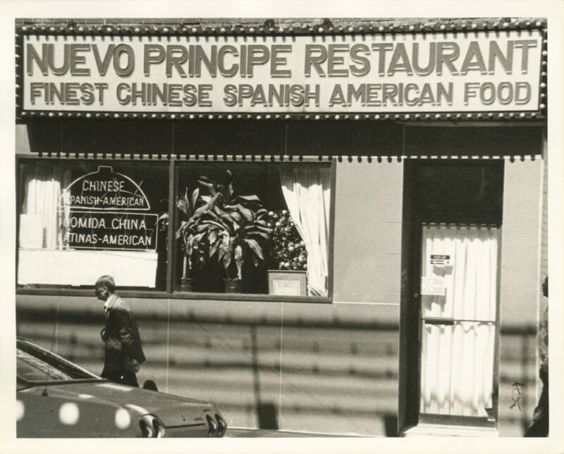 Black-and-white photo of a restaurant with a sign reading "NUEVO PRÍNCIPE RESTAURANT: FINEST CHINESE SPANISH AMERICAN FOOD." A man walks past the window, and a second sign lists food in Chinese, Spanish, and English.