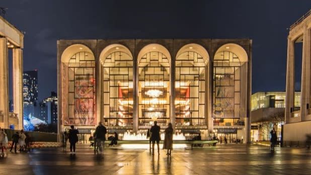Exterior of Lincoln Center brightly lit at night, featuring large arched windows. People stand in front of a glowing fountain, creating a lively atmosphere.