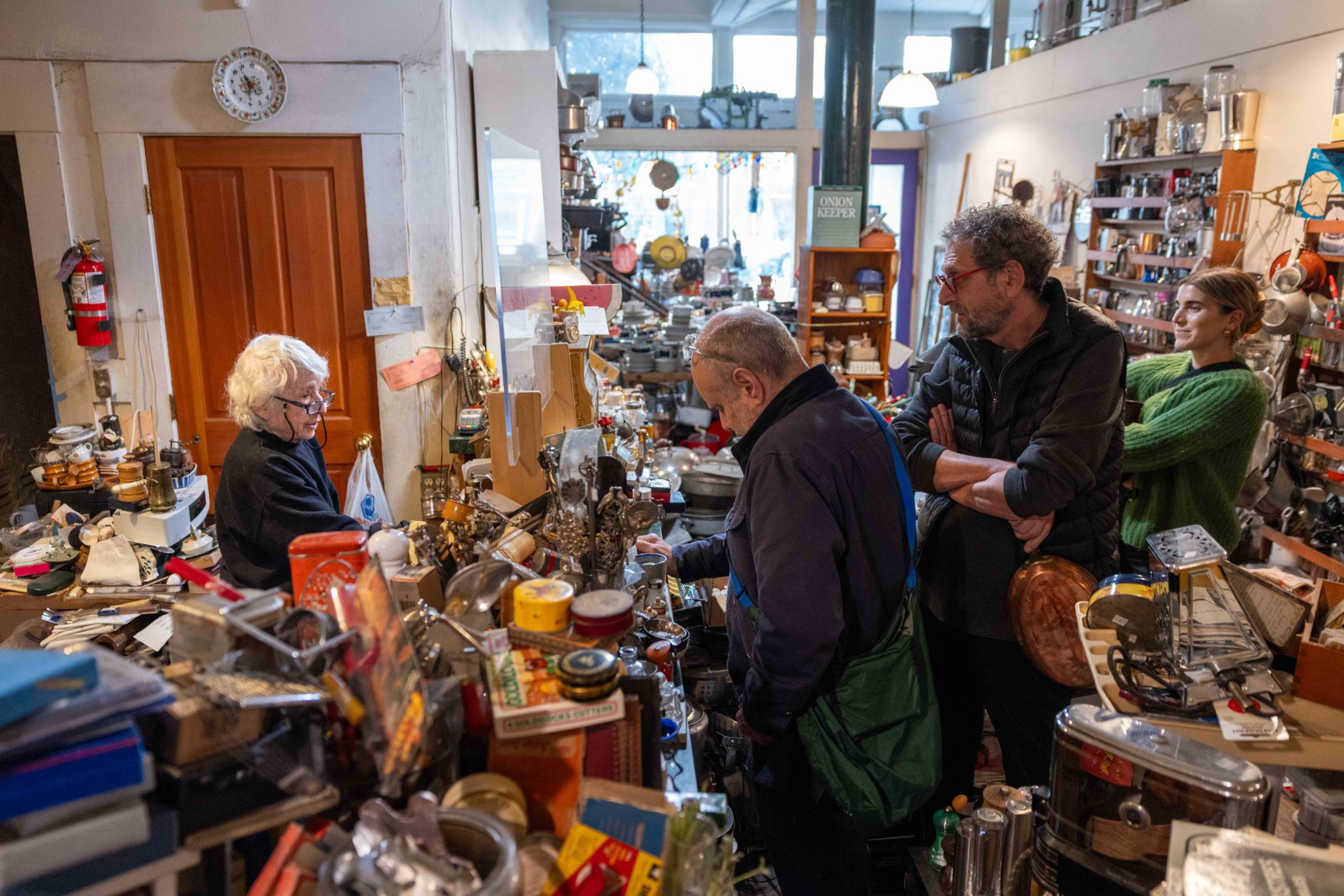 An elderly woman helps three customers in a cluttered antique shop filled with various household items and gadgets.