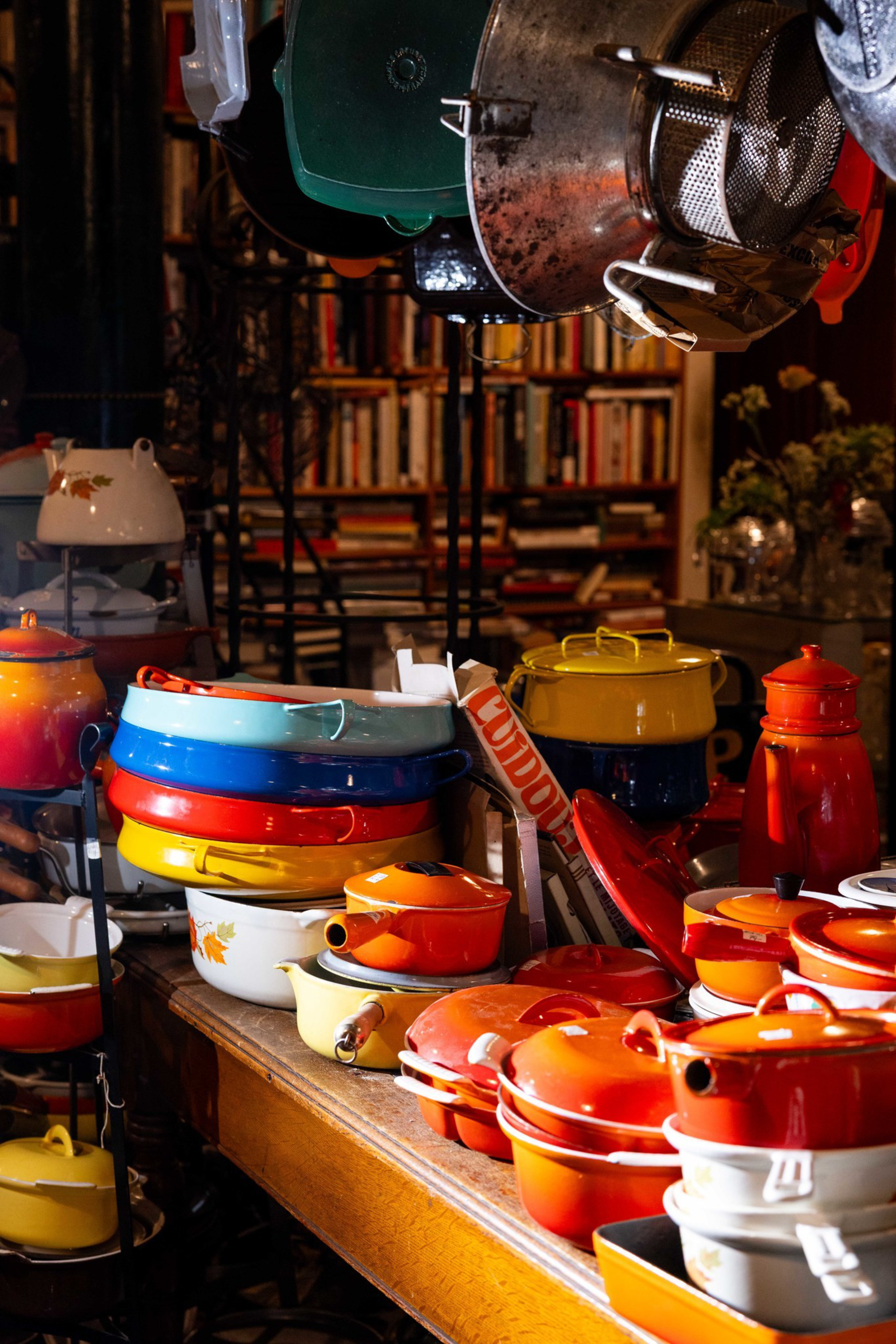 Colorful enamel cookware, including pots and pans in red, orange, yellow, blue, and white, are stacked on a wooden table with bookshelves blurred in the background.