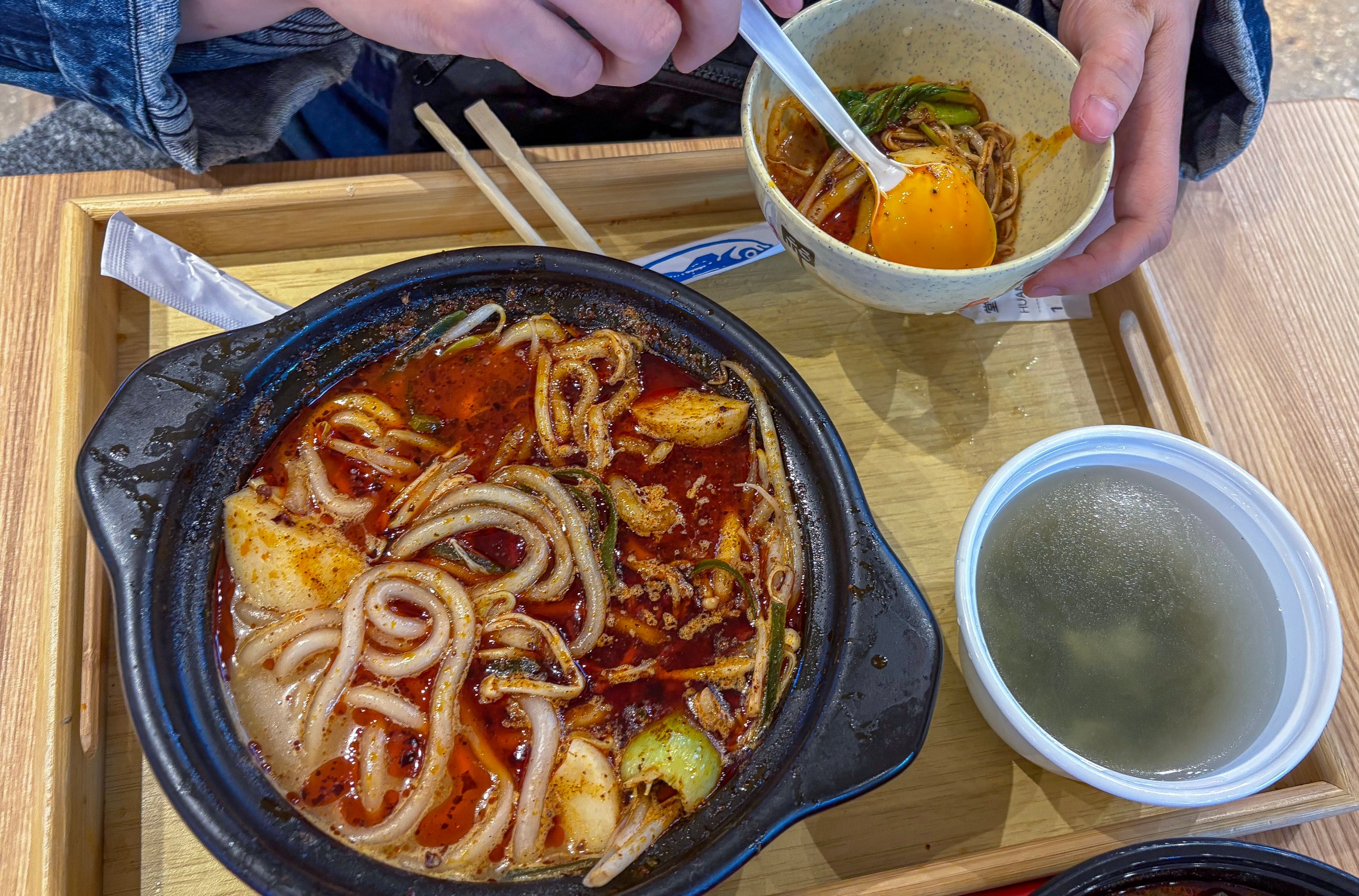 A bowl of broth and noodles on a tray with someone mixing yolk in a separate bowl.