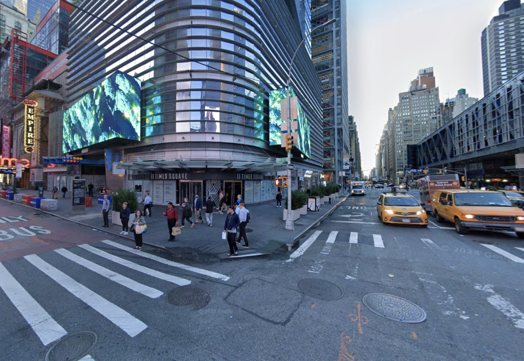 A city street corner with pedestrians walking on the sidewalk and crosswalk, multiple vehicles on the road, and tall buildings lining the street.