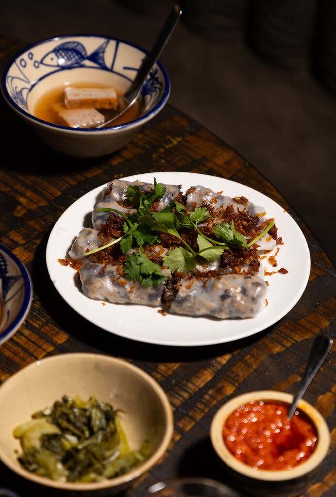 A plate of bánh cuốn, a half-noodle, half-crepe of fermented rice flour, rolled up around ground pork and wood-ear mushrooms and topped with crispy shallots and cilantro, with side saucers of nước chấm, pickles and chile sauce.