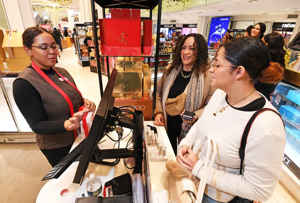 A Macy's employee helping two women with last-minute Christmas shopping at a perfume counter.